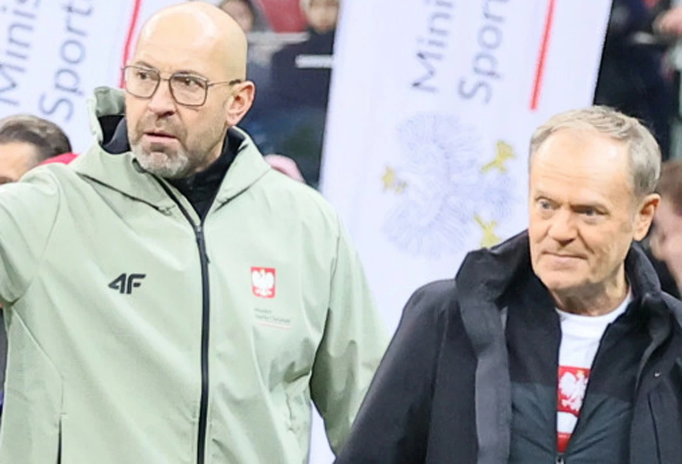 Minister sportu i turystyki Jakub Rutnicki, premier Donald Tusk Dwóch mężczyzn w kurtkach z polskim godłem stoi obok siebie na tle banerów z napisem 'Ministerstwo Sportu', jeden z nich wskazuje ręką na coś poza kadrem.