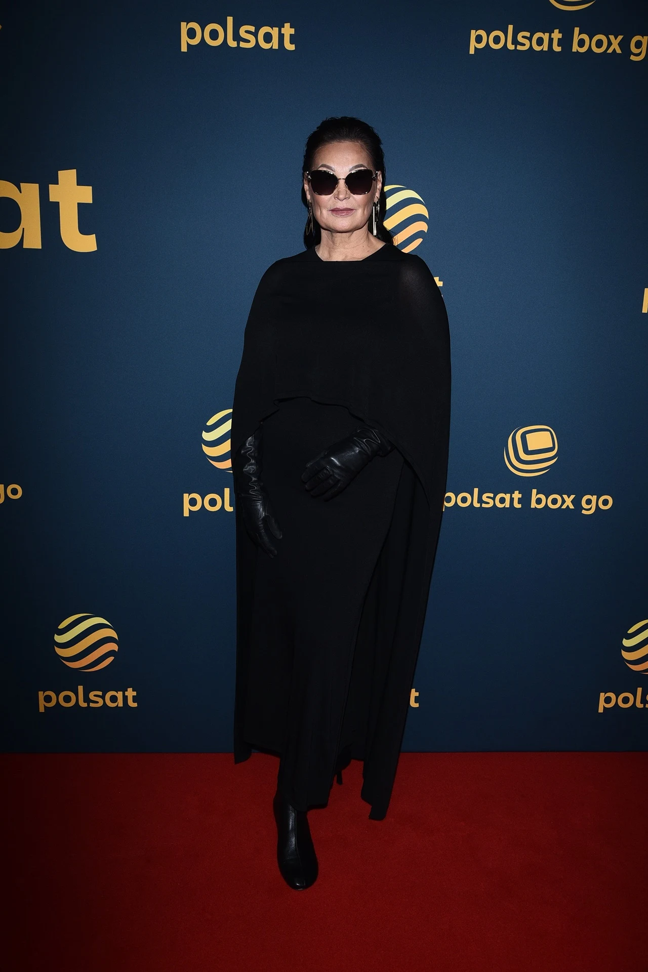 Kobieta w ciemnych okularach, ubrana cała na czarno ze skórzanymi rękawiczkami, stoi na czerwonym dywanie przed ścianką z logo Polsat, tło granatowe z żółtymi napisami.