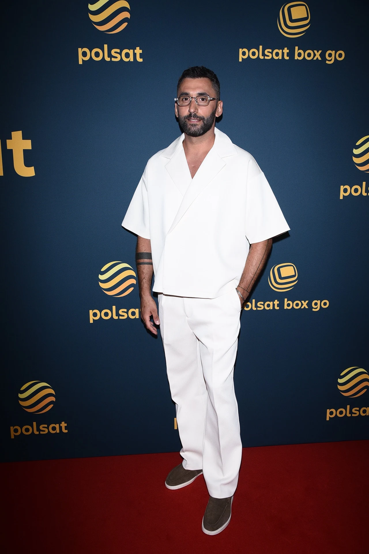 Mężczyzna z ciemnymi włosami i brodą ubrany w białą koszulę o luźnym kroju oraz białe spodnie i szare buty stoi na czerwonym dywanie przed ścianką z logo Polsat oraz Polsat Box Go.