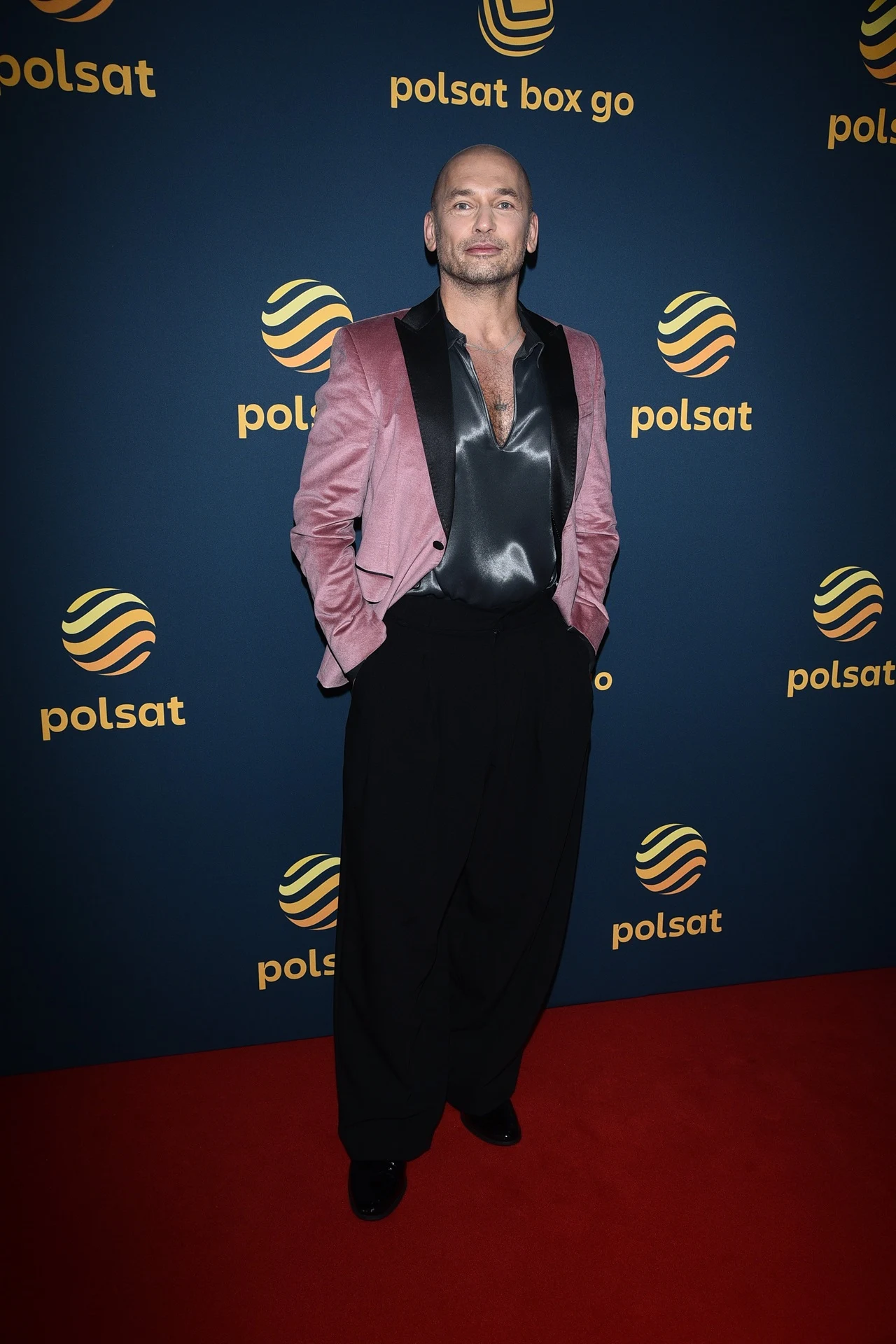 Mężczyzna w różowej marynarce i czarnych, szerokich spodniach pozuje na tle ścianki z logotypami Polsat, stojąc na czerwonym dywanie. Jest elegancko ubrany, z rozpiętą koszulą pod marynarką.