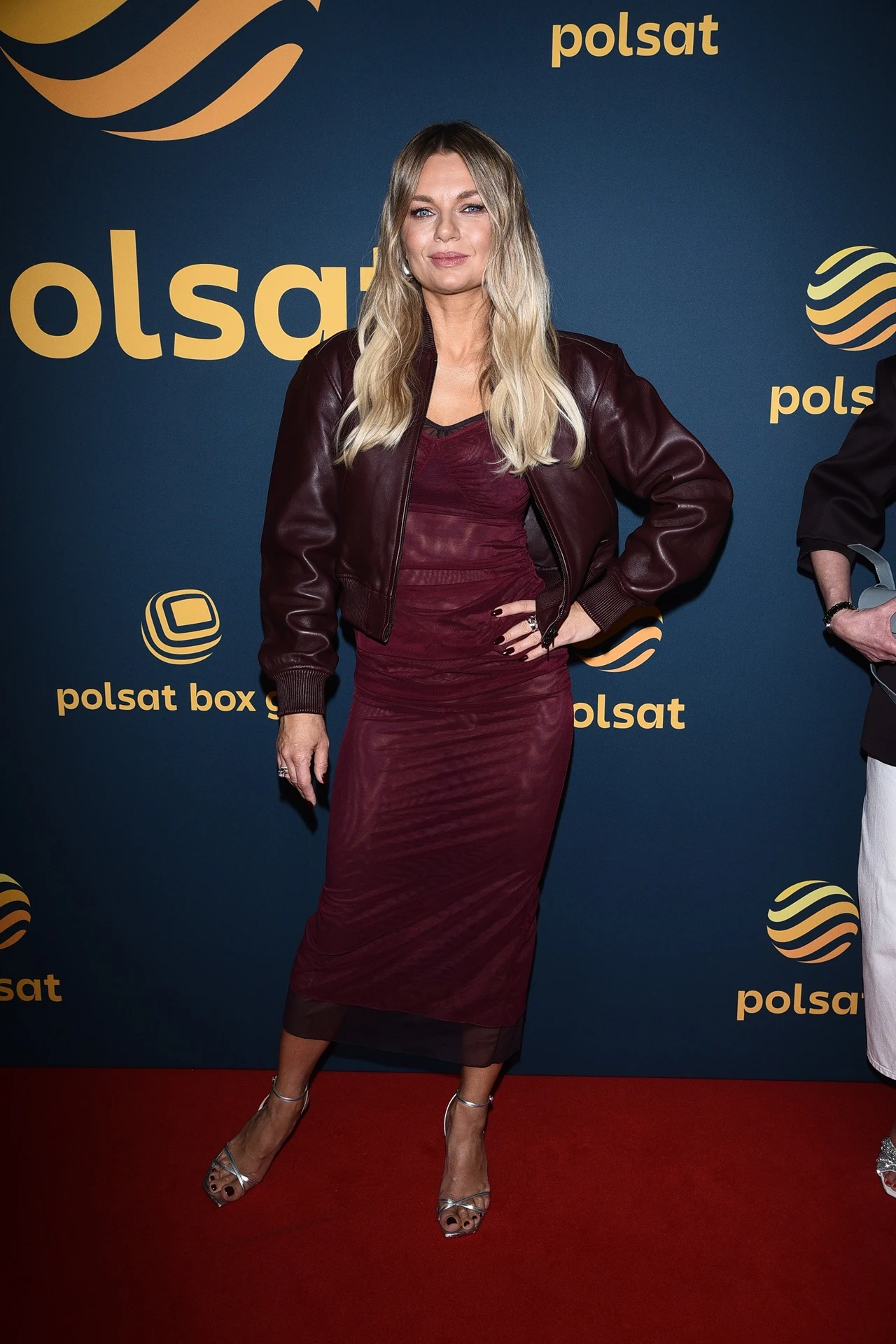 Kobieta w bordowej, dopasowanej sukience i skórzanej kurtce stoi na czerwonym dywanie na tle ścianki z logiem Polsat, pozując do zdjęcia.