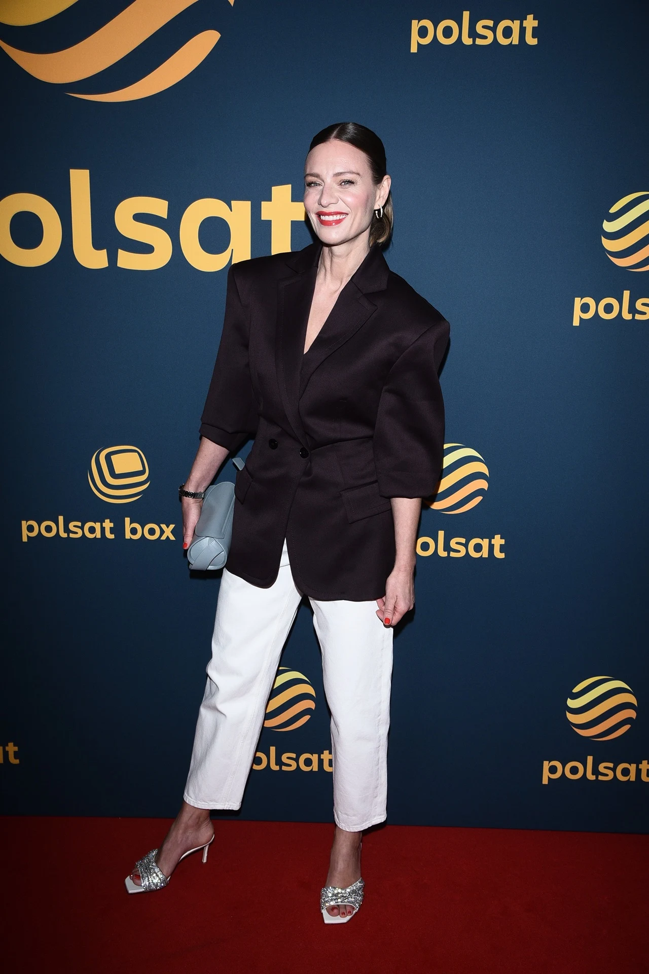 Elegancko ubrana kobieta pozuje na tle granatowej ścianki z żółtym logo Polsat, ubrana w czarną marynarkę, białe spodnie i srebrne buty, trzyma srebrną kopertówkę.