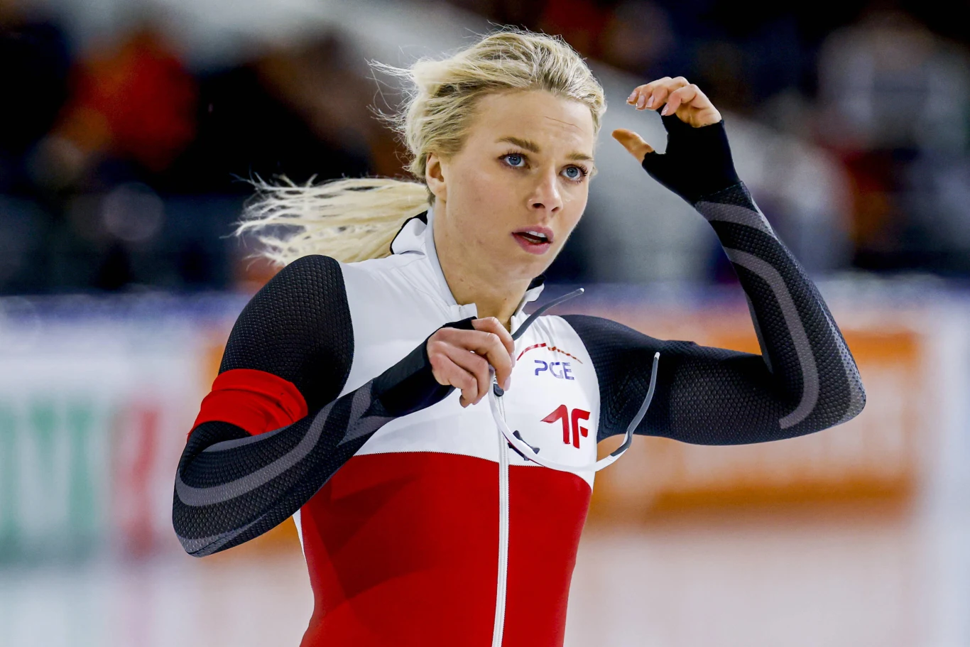 Zawodniczka w stroju sportowym na torze łyżwiarskim, ubrana w kombinezon w barwach narodowych z widocznym logo sponsora, skoncentrowana po przejeździe.