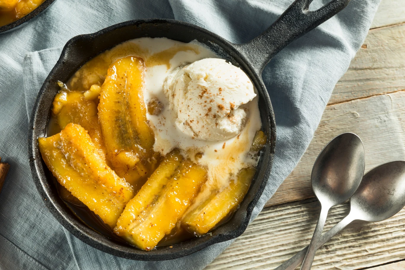 Karmelizowane banany w małej żeliwnej patelni z kulką waniliowych lodów, całość podana na niebieskiej serwetce, obok dwie łyżki.