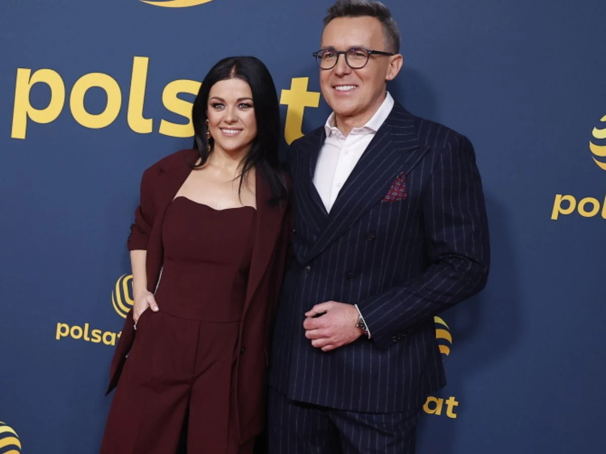 Kobieta w bordowym garniturze i mężczyzna w granatowym garniturze z białą koszulą stoją razem na tle ściany z logotypami Polsat, oboje uśmiechają się do obiektywu.