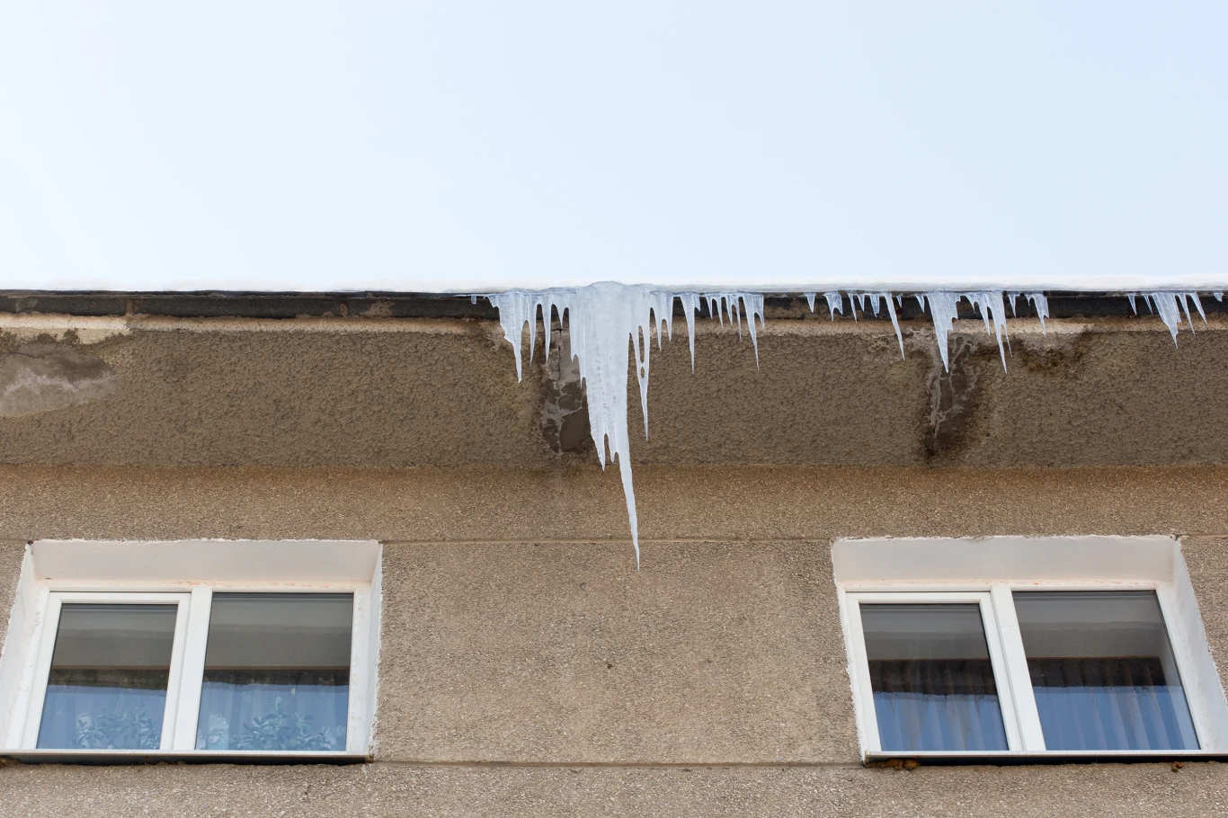 Zimą zwisające sople z balkonów i dachów stają się realnym zagrożeniem dla przechodniów. Sople lodu zwisające z krawędzi dachu budynku mieszkalnego, poniżej dwa okna z białymi ramami, ściana o jasnym kolorze.
