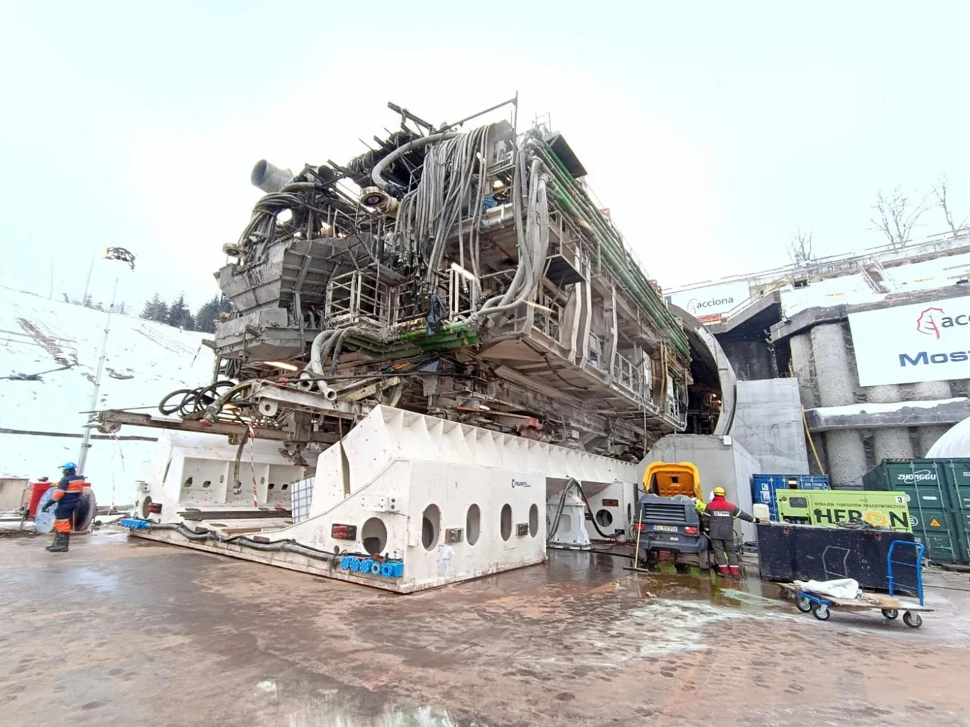 Obrócenie maszyny TBM na budowie S19 to trudna operacja Duża maszyna drążąca tunel z licznymi przewodami i elementami technicznymi, ustawiona na placu budowy w otoczeniu sprzętu, narzędzi oraz kilku pracowników w odzieży ochronnej. W tle infrastruktura budowlana i lekko zaśnieżony teren.