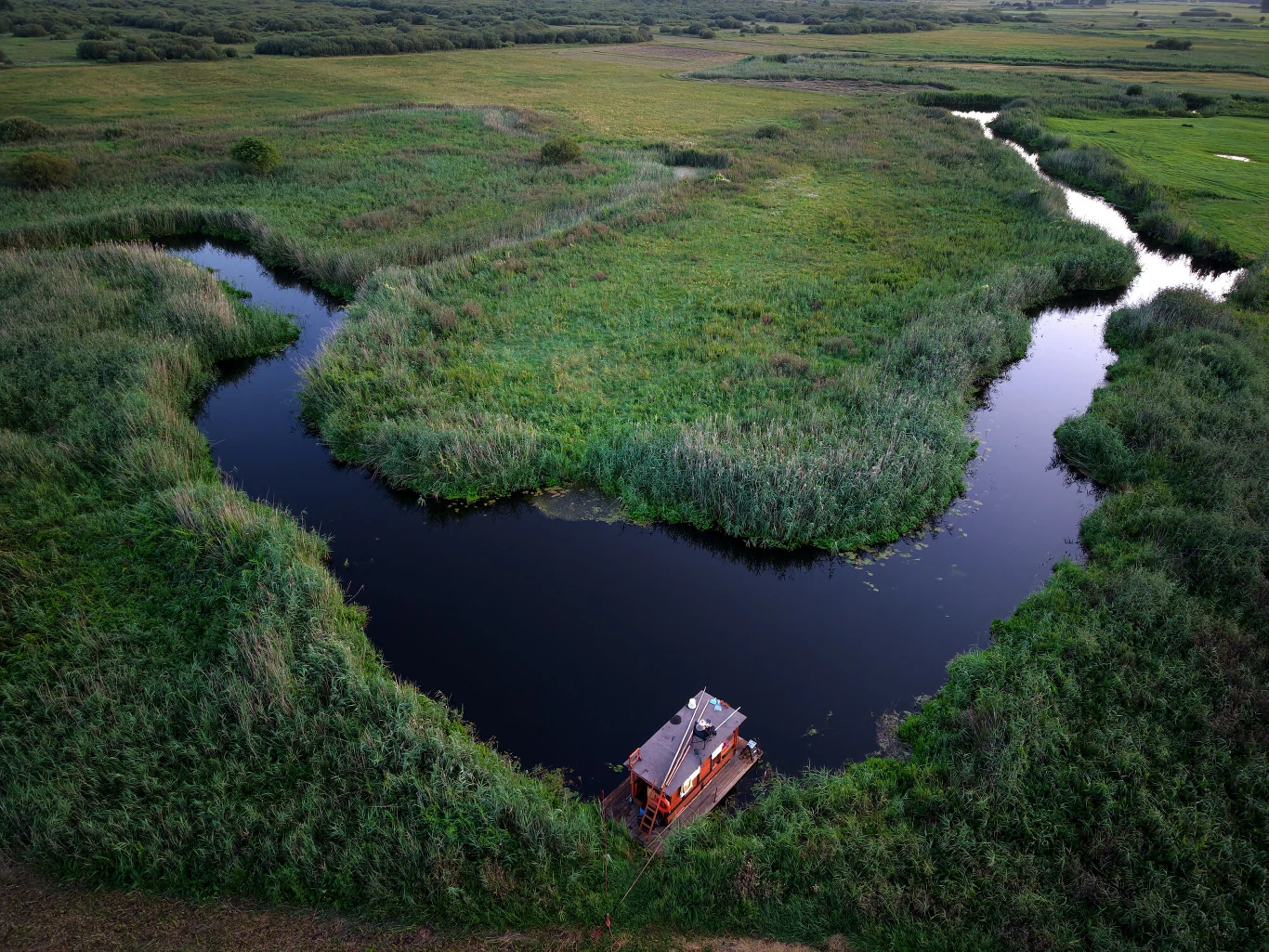 Drewniana altana na wodzie przy malowniczo meandrującej rzece, otoczona gęstą zielenią trzcin i łąk, z szeroką perspektywą rozlewisk i pól na horyzoncie.
