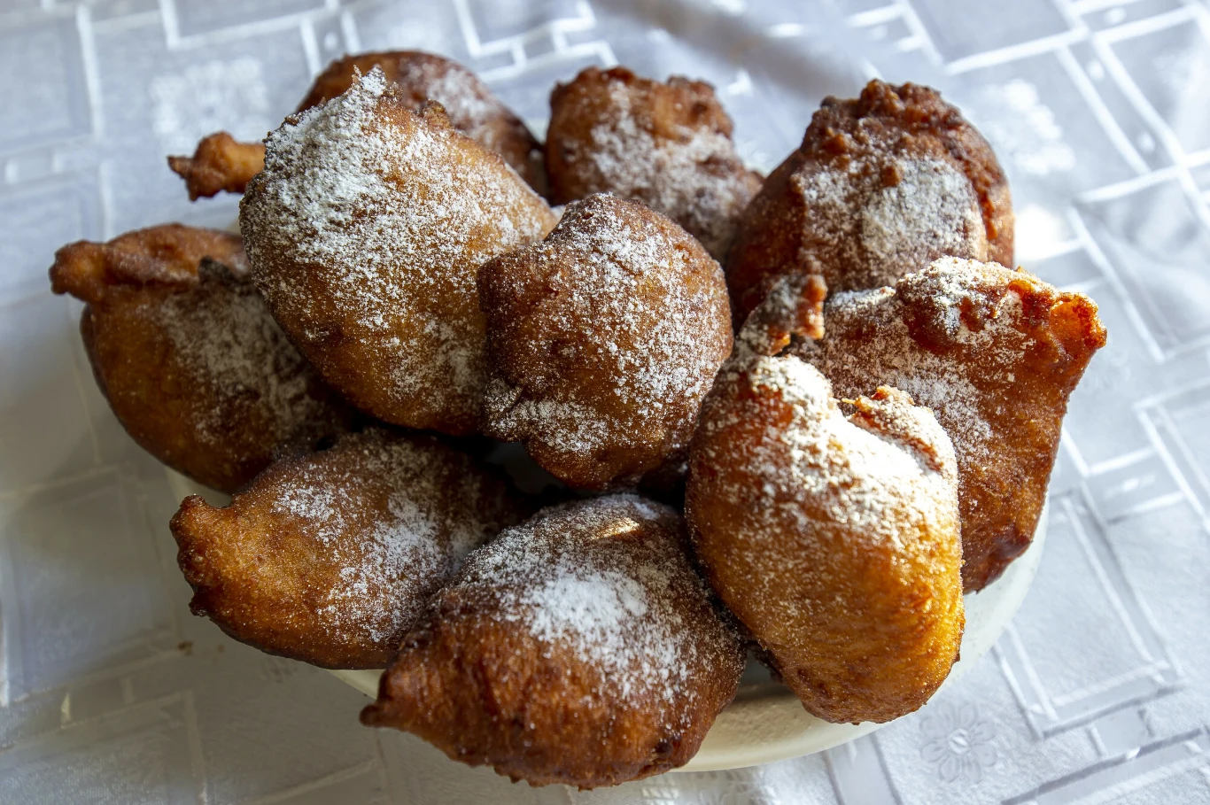 Stos pączków posypanych cukrem pudrem na białym talerzu ustawionym na jasnym, wzorzystym obrusie.