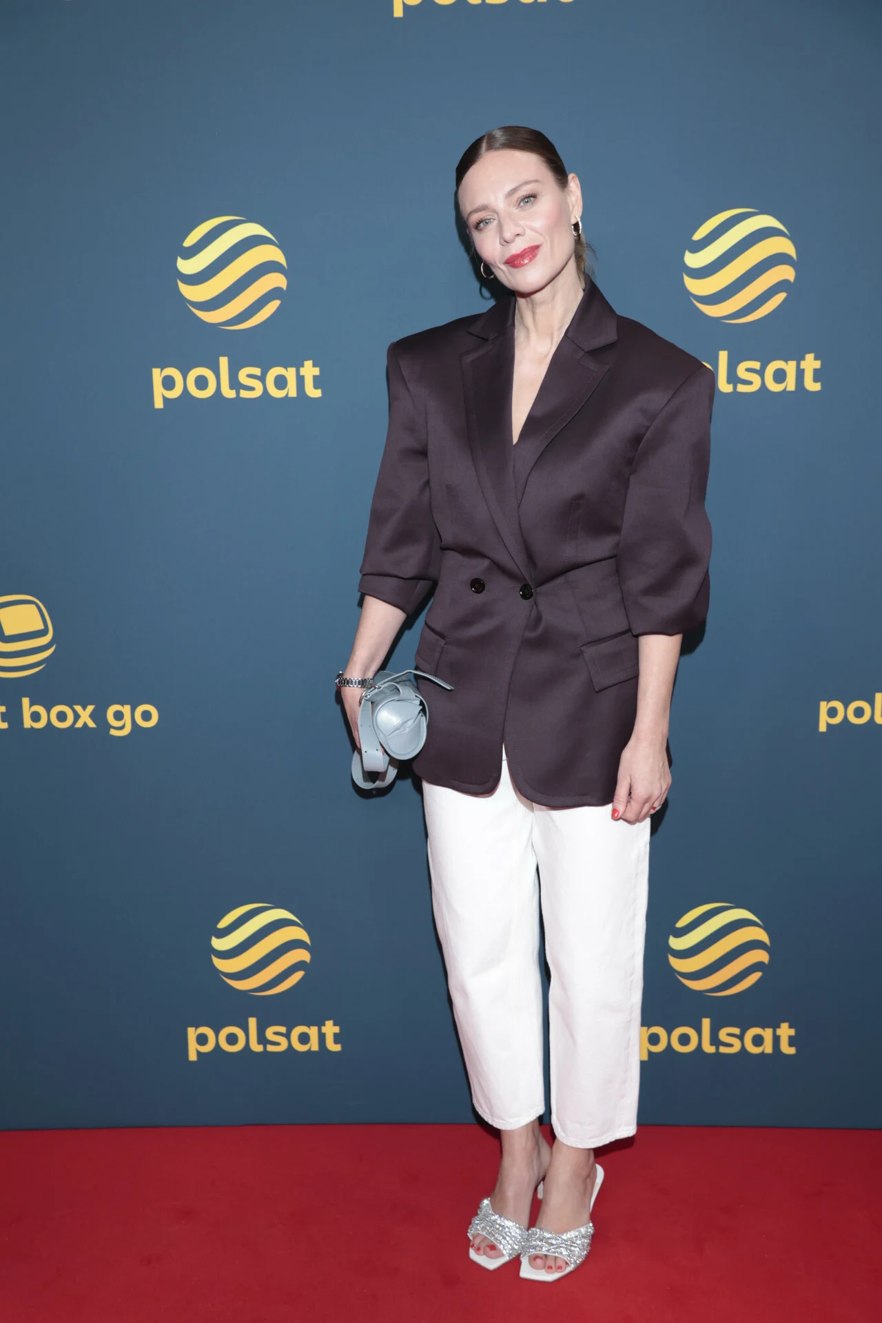 Kobieta stojąca na czerwonym dywanie, ubrana w czarną marynarkę z szerokimi ramionami, białe spodnie oraz srebrne buty na obcasie. W dłoni trzyma szarą torebkę. W tle widoczne logo Polsat na ciemnoniebieskiej ścianie.