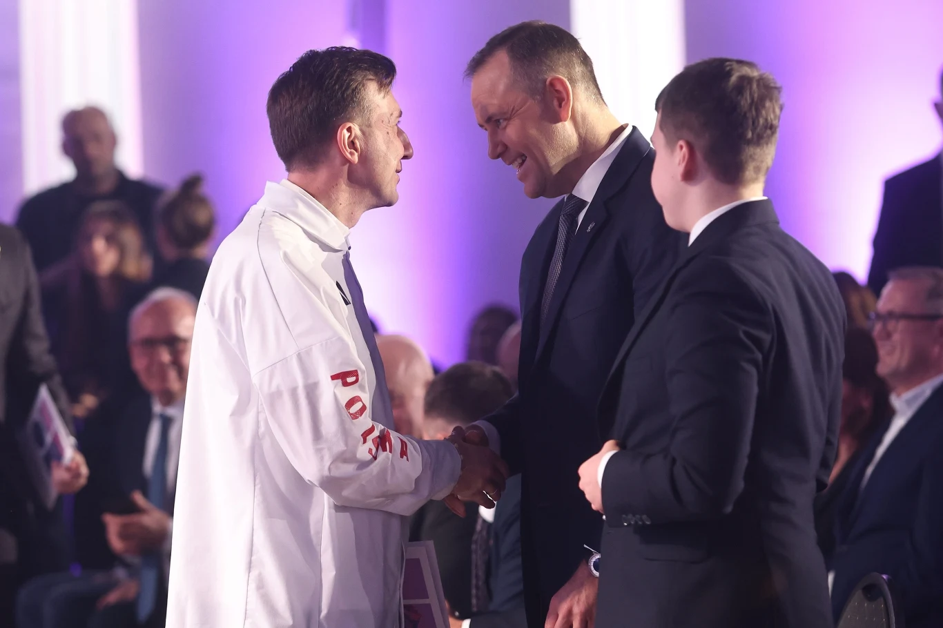 Dwie osoby w oficjalnych strojach uśmiechają się i wymieniają uścisk dłoni podczas formalnego wydarzenia, w tle widoczni są siedzący uczestnicy w garniturach, całość oświetlona jest fioletowym światłem.