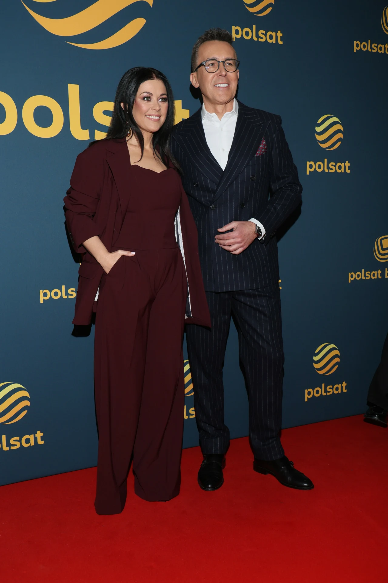 Kobieta w bordowym kombinezonie i mężczyzna w ciemnym garniturze stoją razem na czerwonym dywanie, uśmiechając się do zdjęcia na tle ścianki z logo Polsat.