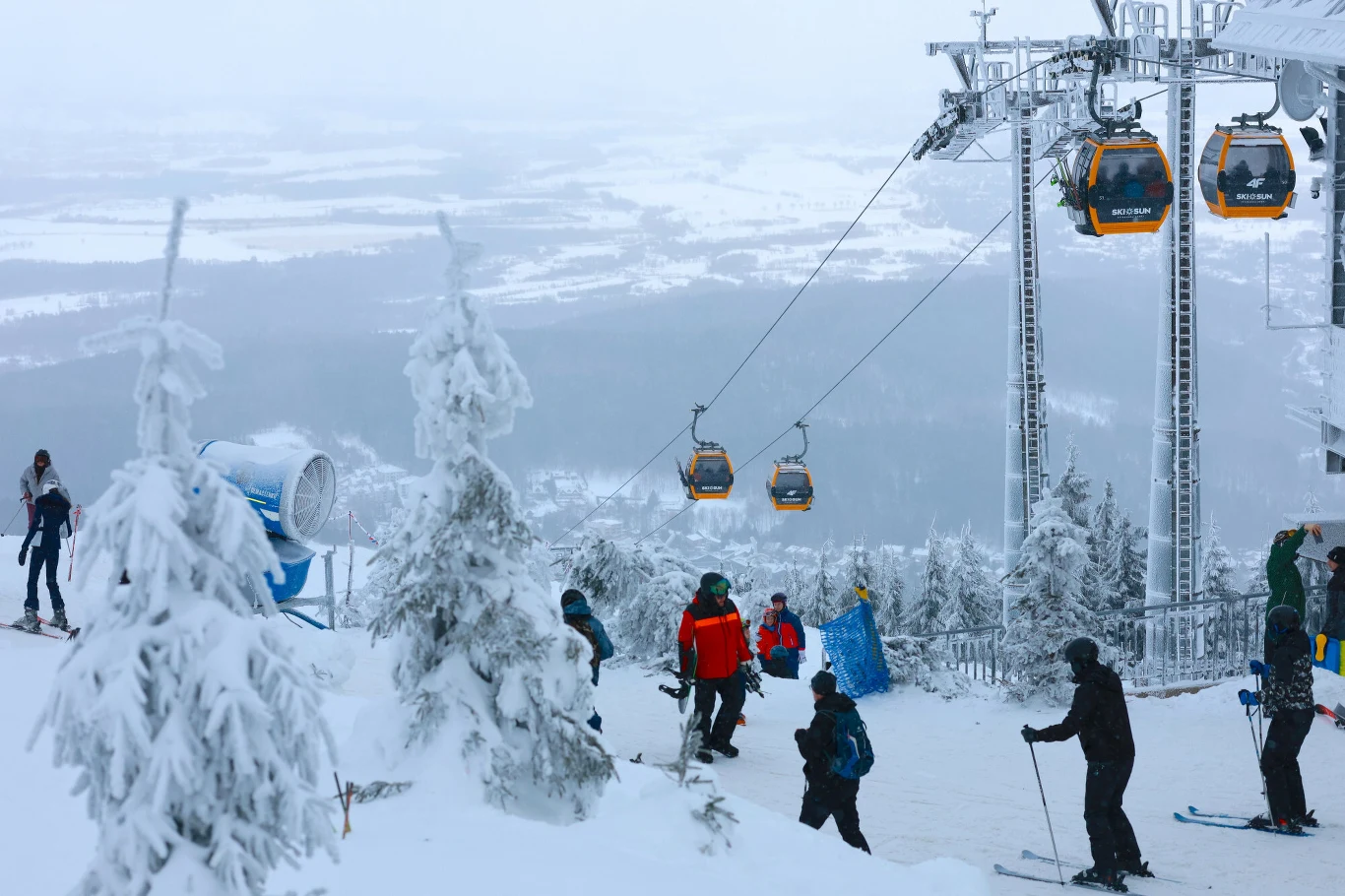 Narciarze i snowboardziści na ośnieżonym stoku górskim w otoczeniu zaśnieżonych drzew, w tle widoczne gondole wyciągu narciarskiego oraz rozległy, zamglony krajobraz doliny.