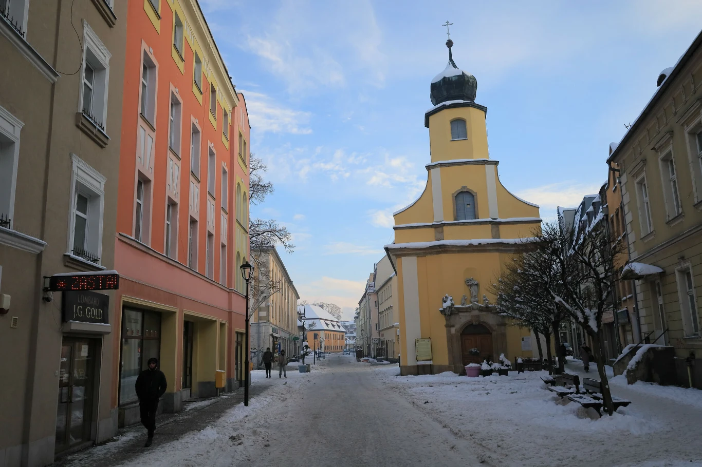 Ośnieżona ulica prowadząca przez centrum miasta, po lewej stronie kolorowa kamienica, po prawej żółty kościół z charakterystyczną wieżą i krzyżem, kilka osób spacerujących, drzewa pokryte śniegiem.