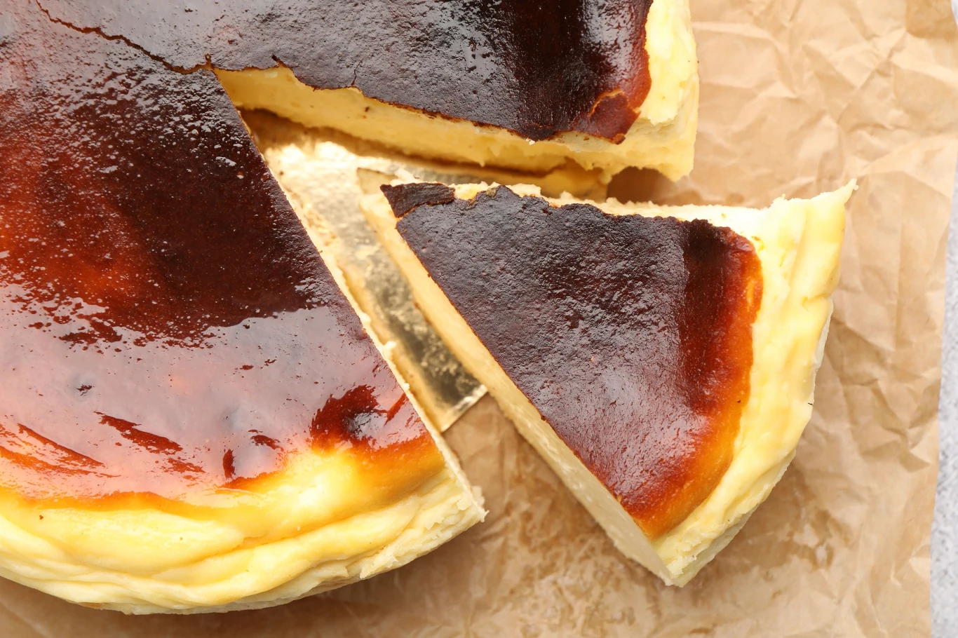 Przygotowania sernika baskijskiego jest banalne. Zrobisz go w mgnieniu oka Sernik baskijski o ciemnej, przypieczonej górze, leżący na arkuszu papieru, z wyciętym trójkątnym kawałkiem odłożonym na bok.