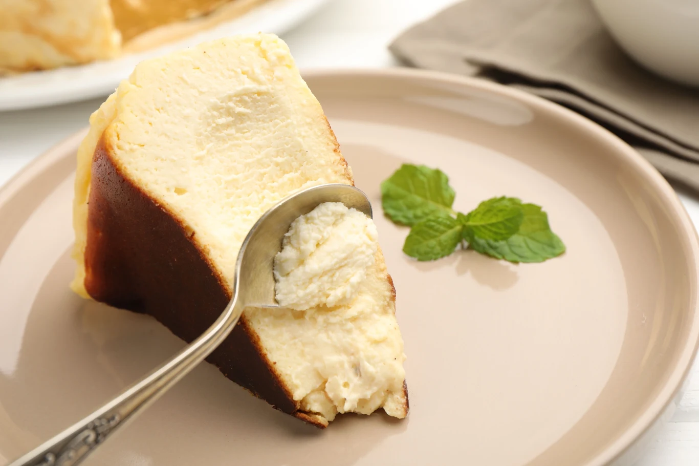 Sernik baskijski jest lekki i kremowy, z przypieczonym wierzchem Kawałek sernika z brązową, wypieczoną skórką, nakłuwany łyżeczką, obok dekoracja z liści mięty na jasnobeżowym talerzu.