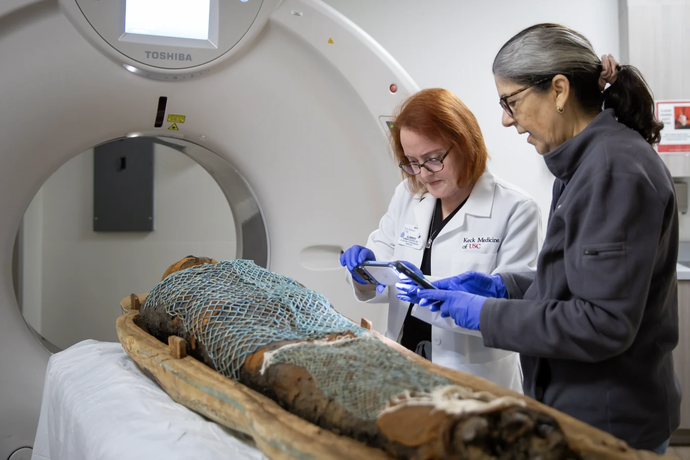 Dwie kobiety w białych fartuchach i rękawiczkach niebieskich analizują starożytną mumię, która leży na stole obok tomografu komputerowego. Jedna z nich używa tabletu, druga obserwuje uważnie procedurę.