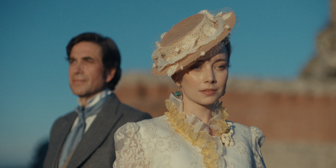 "Lalka" film z 2026 roku Kobieta w stroju z epoki, ozdobionym kapeluszem i falbanami, z poważnym wyrazem twarzy stoi na pierwszym planie. W tle mężczyzna w eleganckim garniturze z muszką. Oboje na tle murów w jasnym świetle dziennym.