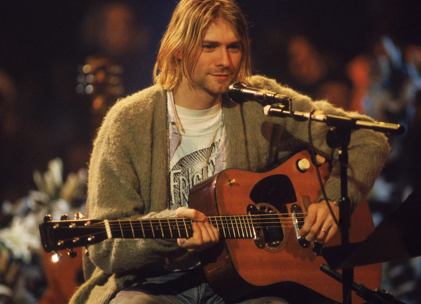 Kurt Cobain nie spodziewał się tak dużego sukcesu Mężczyzna z długimi blond włosami siedzi na scenie, trzymając gitarę akustyczną. Ubrany w szary sweter i jasną koszulkę. Gra na gitarze i śpiewa do mikrofonu, scena oświetlona jest miękkim światłem, a w tle widoczna jest niewyraźna publiczność.