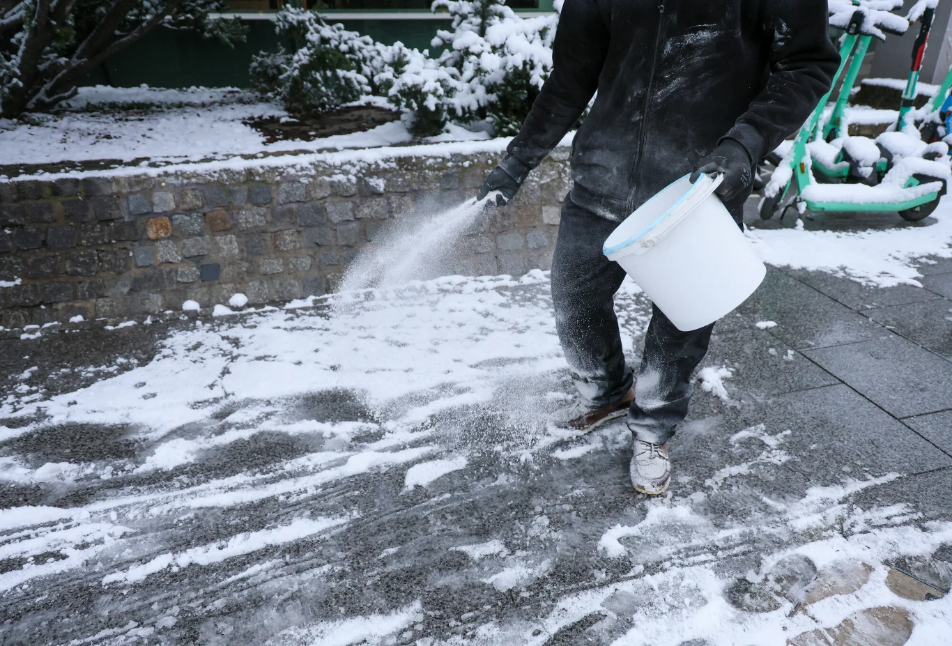 W Niemczech coraz popularniejsze są zakazy soli drogowej Osoba ubrana w ciemną kurtkę i spodnie posypuje ośnieżony chodnik solą z białego wiadra. W tle widoczny kamienny mur, przysypane śniegiem krzewy oraz zielony sprzęt miejskiej wypożyczalni.
