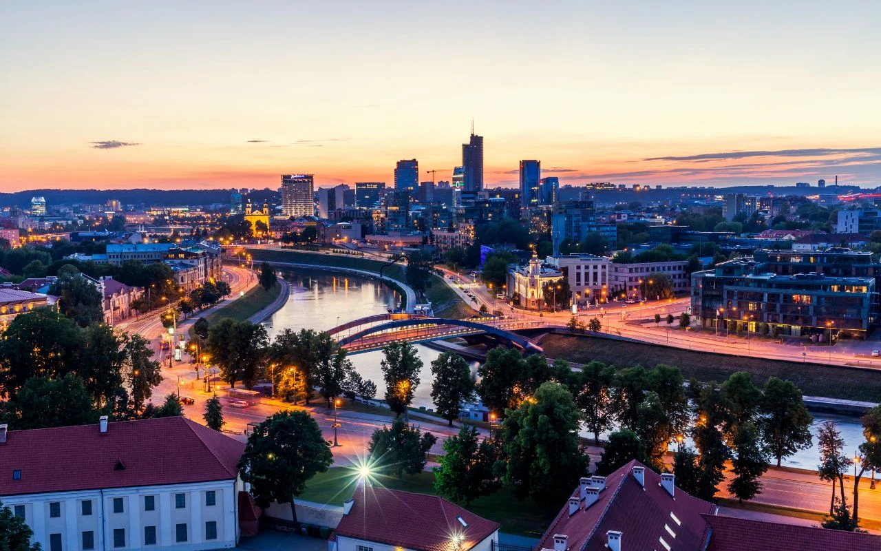 Panorama współczesnego miasta z nowoczesnymi wieżowcami, starą zabudową, rzeką płynącą przez środek oraz mostami i zielenią. Miasto oświetlone światłami ulicznymi podczas zachodu słońca.