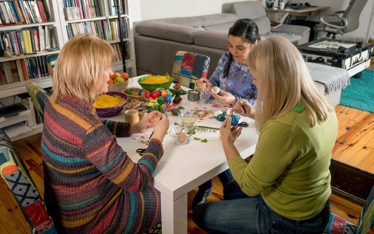 Trzy kobiety siedzą przy stole, ozdabiają pisanki w otoczeniu różnokolorowych materiałów, warzyw i jajek, w tle widać regał z książkami i fragment salonu.