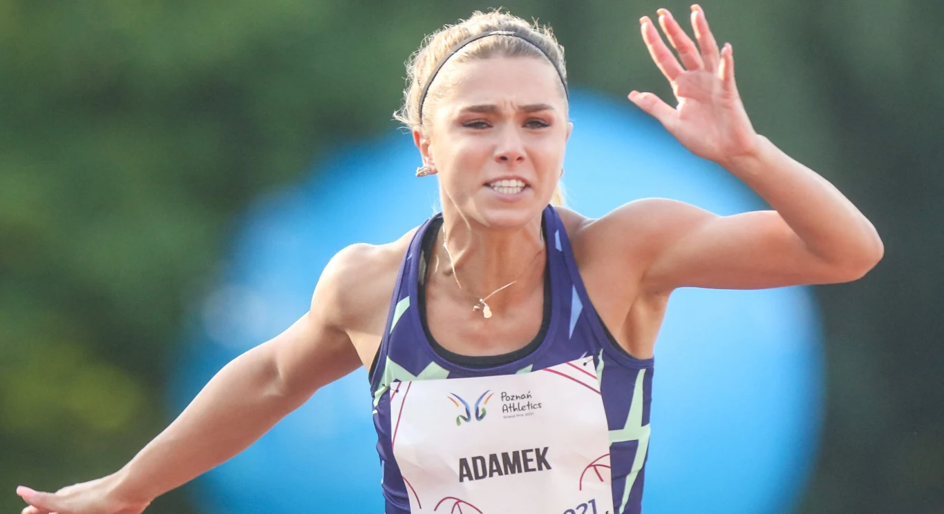 Kobieta w sportowym stroju biegnąca na bieżni, wyraźnie skoncentrowana na wysiłku fizycznym, z widocznym numerem startowym i napisem 'ADAMEK' na piersi.