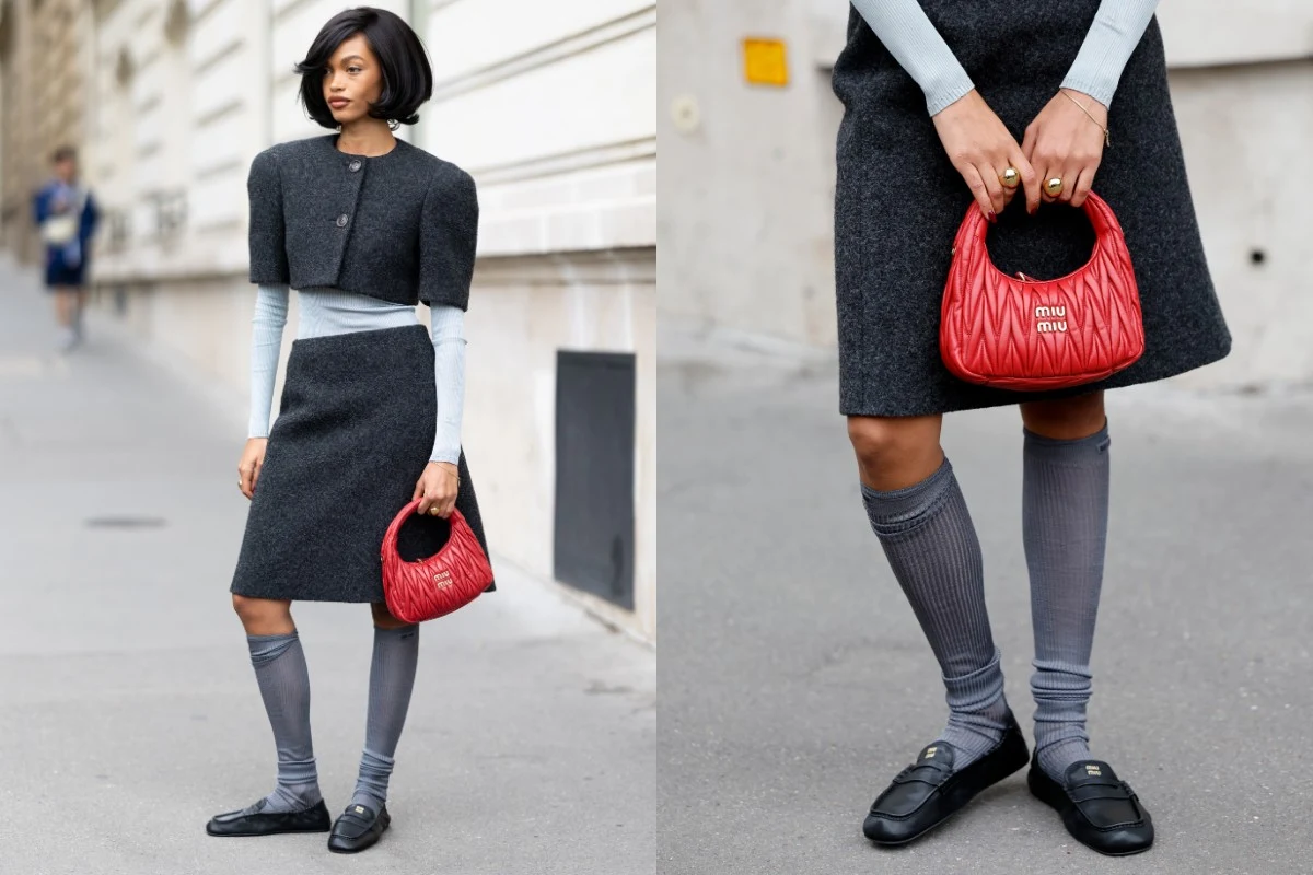 Modne buty na wiosnę 2026 Kobieta ubrana w grafitowy komplet składający się z krótkiej pelerynki i spódnicy, jasnoszary cienki golf, szare podkolanówki oraz czarne mokasyny, trzyma w dłoni małą czerwoną pikowaną torebkę. Scena miejska w tle.