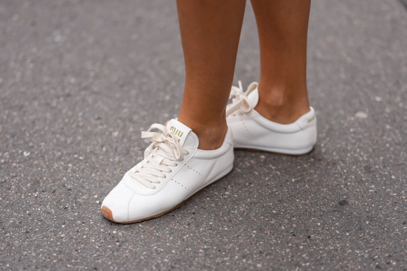 Najmodniejsze sneakersy na wiosnę Białe sneakersy marki Miu Miu na stopach osoby stojącej na szarej asfaltowej nawierzchni, z widocznymi nogami od kostek w dół.