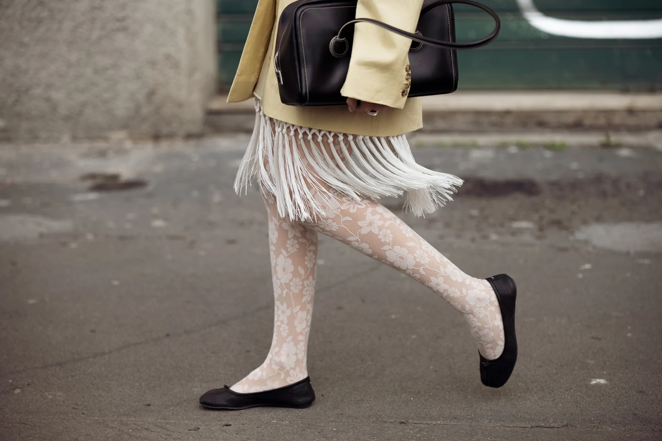 Jakie buty wybrać na wiosnę? Dolna część ciała osoby idącej chodnikiem, ubrane w białe rajstopy w kwiatowy wzór, jasną spódnicę z frędzlami oraz czarne baleriny. Osoba trzyma czarną torebkę.