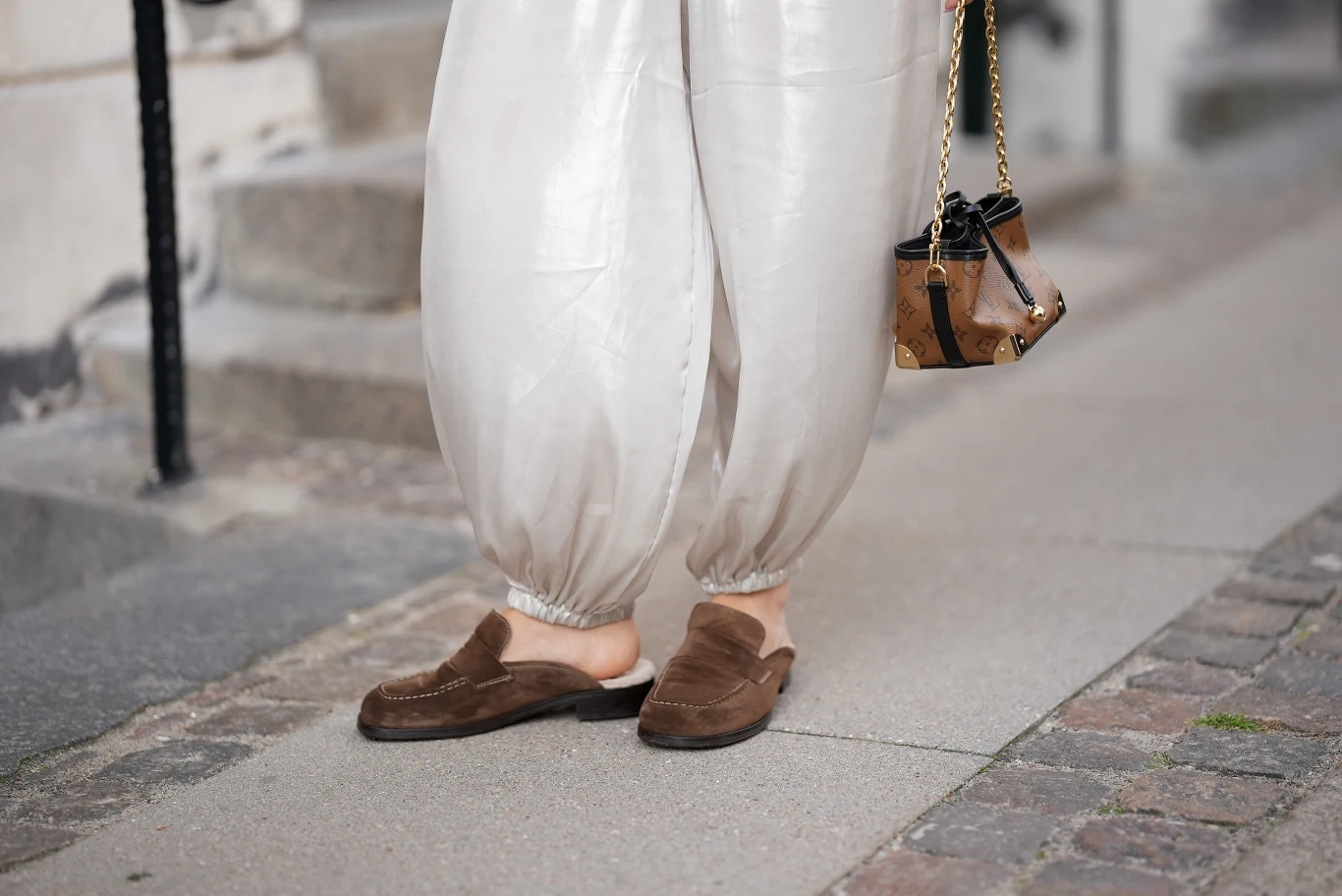 Wygodne buty na wiosnę 2026 Jasne, satynowe spodnie z gumką w nogawkach, brązowe zamszowe klapki na stopach oraz mała torebka na łańcuszku trzymana w ręce, sceneria miejska z chodnikiem i schodkami w tle.
