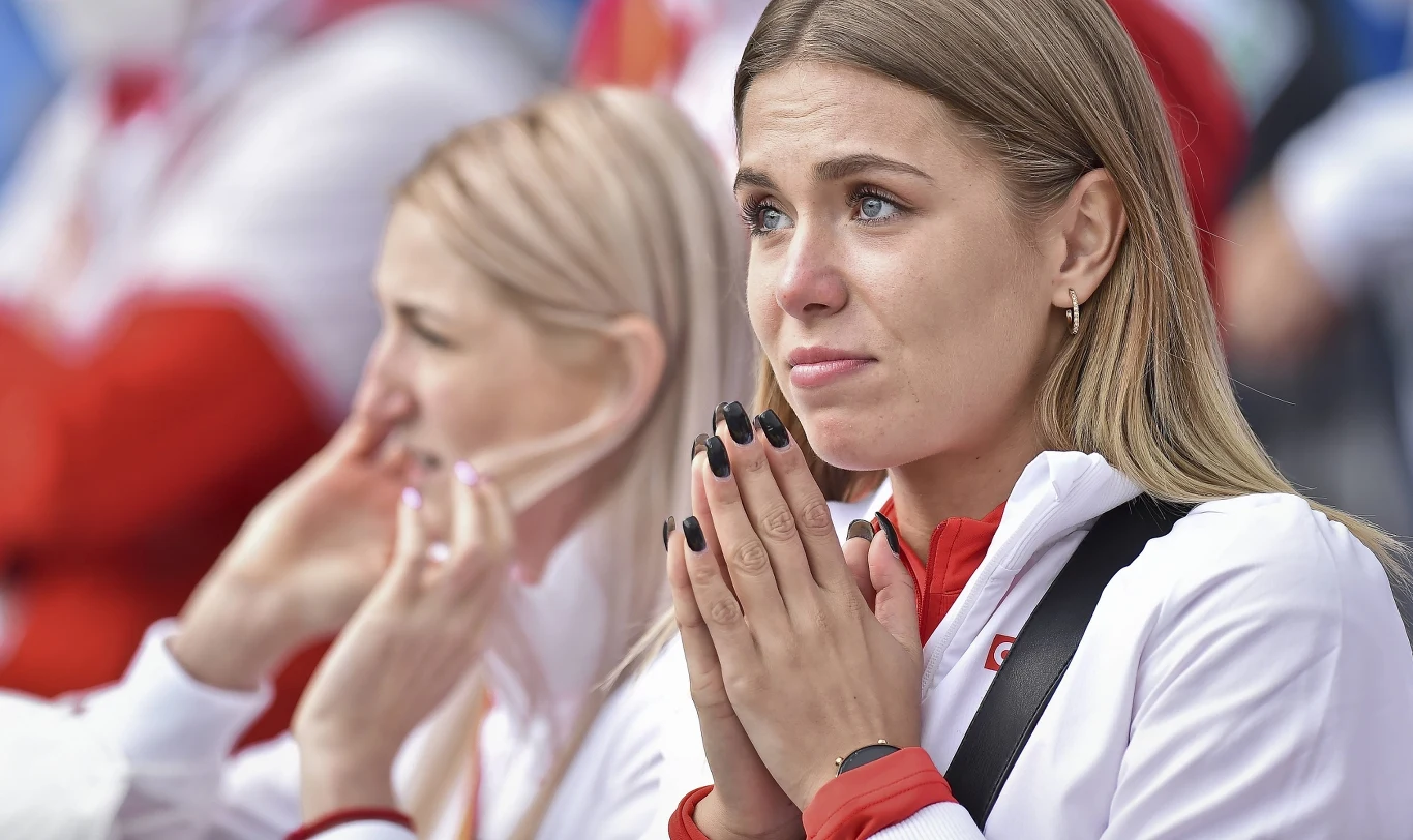 Klaudia Adamek Kobieta o jasnych włosach trzyma dłonie złożone w geście modlitwy lub skupienia, ma wyraz twarzy pełen napięcia i emocji, w tle widoczna jest druga kobieta również skupiona na wydarzeniu.