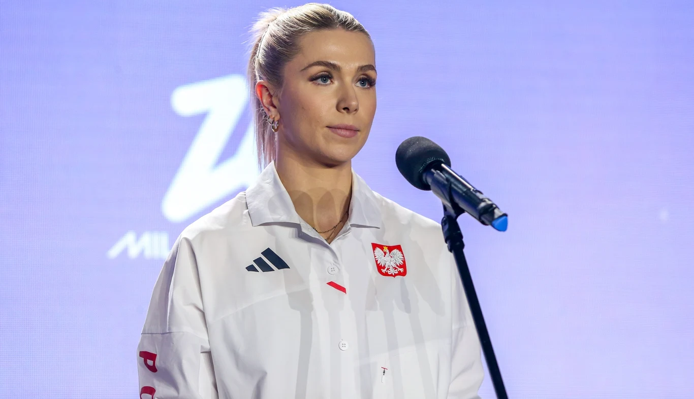Klaudia Adamek Kobieta w białej koszuli z polskim godłem stoi przy mikrofonie na tle niebieskiego ekranu z rozmytym napisem. Blond włosy związane w kok, delikatny makijaż, poważny wyraz twarzy.