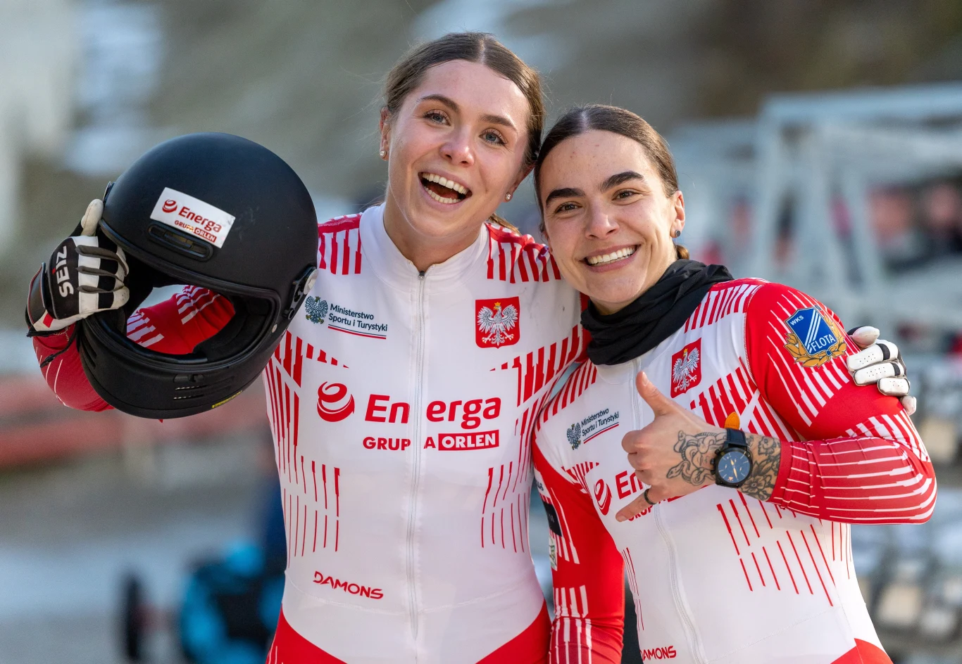 Klaudia Adamek i Linda Weiszewski, czyli TEAM Dwie uśmiechnięte zawodniczki w sportowych kombinezonach z logotypami sponsorów, w barwach Polski. Jedna trzyma kask, druga pokazuje gest kciuka w górę. W tle rozmyty fragment toru bobslejowego.