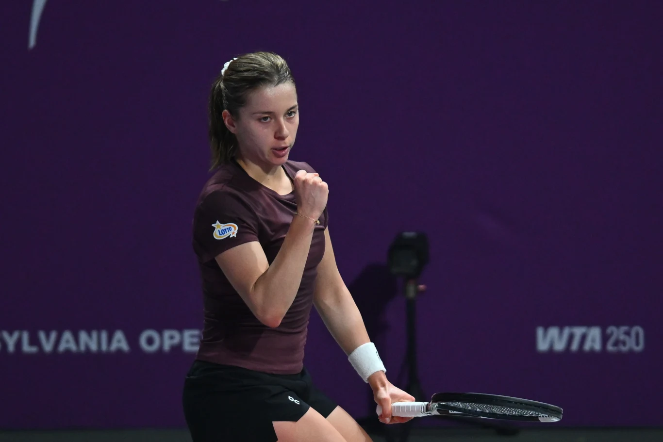 Tenisistka z uniesioną pięścią w geście triumfu, trzymająca rakietę na tle fioletowego kortu z widocznymi logotypami turnieju.