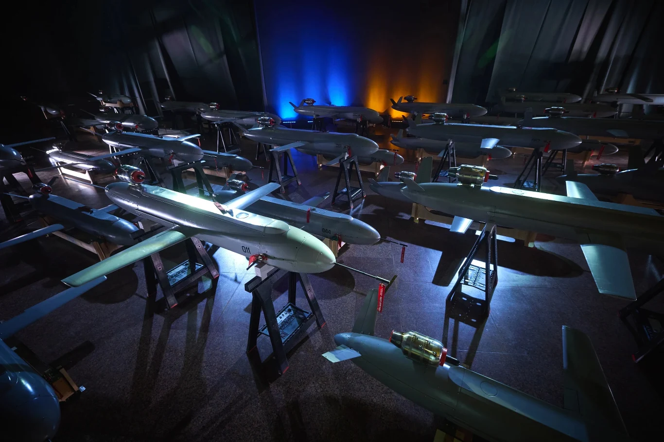 Modele wojskowych dronów rakietowych ustawione na stojakach w pomieszczeniu z ciemnym tłem i kolorowym oświetleniem, głównie niebieskim oraz pomarańczowym.