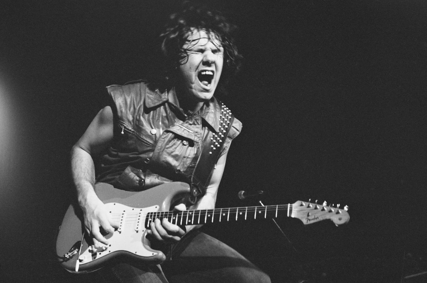 Gary Moore podczas występu w Osace (Japonia), 25 lutego 1984 r. Mężczyzna grający energicznie na gitarze elektrycznej, ubrany w skórzaną kamizelkę, z wyrazistą ekspresją twarzy wyrażającą emocje związane z muzyką, oświetlony światłem scenicznym na ciemnym tle.