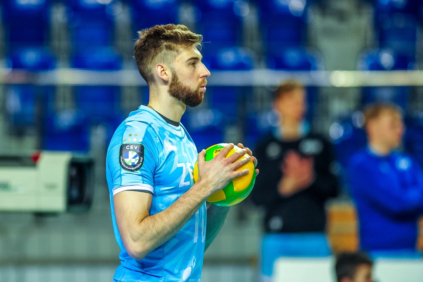 Siatkarz w niebieskim stroju trzyma żółto-zieloną piłkę, przygotowując się do zagrywki, w tle rozmyci kibice oraz niebieskie krzesełka na trybunach.