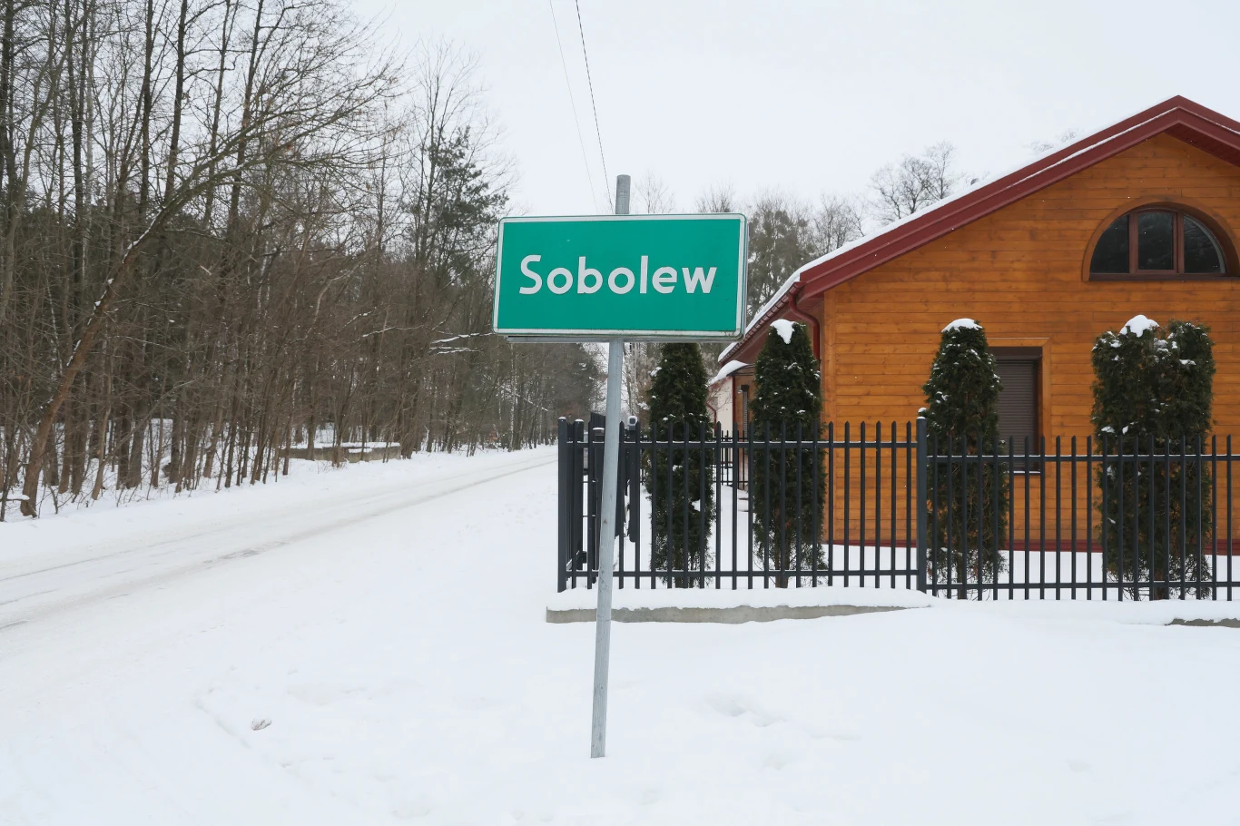 Tablica z nazwą miejscowości Sobolew ustawiona przy ośnieżonej drodze, w tle drewniany budynek i rząd przyciętych tujek, zimowy krajobraz i drzewa wokół.