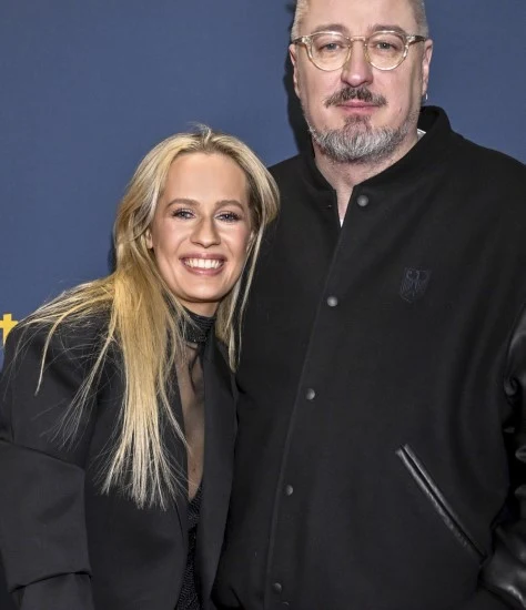 Dwie osoby stojące obok siebie na tle niebieskiej ściany z żółtym napisem. Kobieta z długimi blond włosami uśmiecha się, a mężczyzna ma okulary i krótką fryzurę. Oboje mają na sobie elegancką, ciemną odzież.