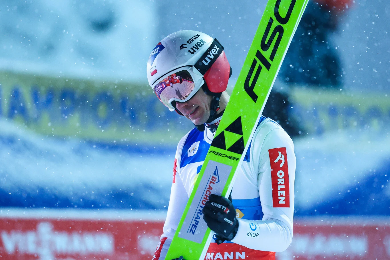Zawodnik w stroju narciarskim oraz kasku trzyma narty z napisem Fischer, na tle padającego śniegu i rozmytego baneru z napisem. Zawodnik wydaje się być zamyślony lub zmęczony po odbytym skoku.