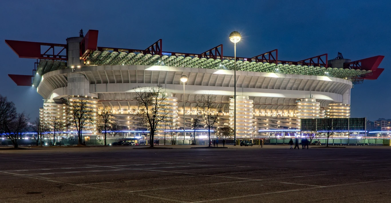 Stadio San Siro. To tutaj w piątkowy wieczór odbędzie się ceremonia otwarcia igrzysk Nowoczesny stadion sportowy oświetlony wieczorem, z widoczną trybuną i elementami konstrukcji architektonicznej, otoczony drzewami i pustym parkingiem.