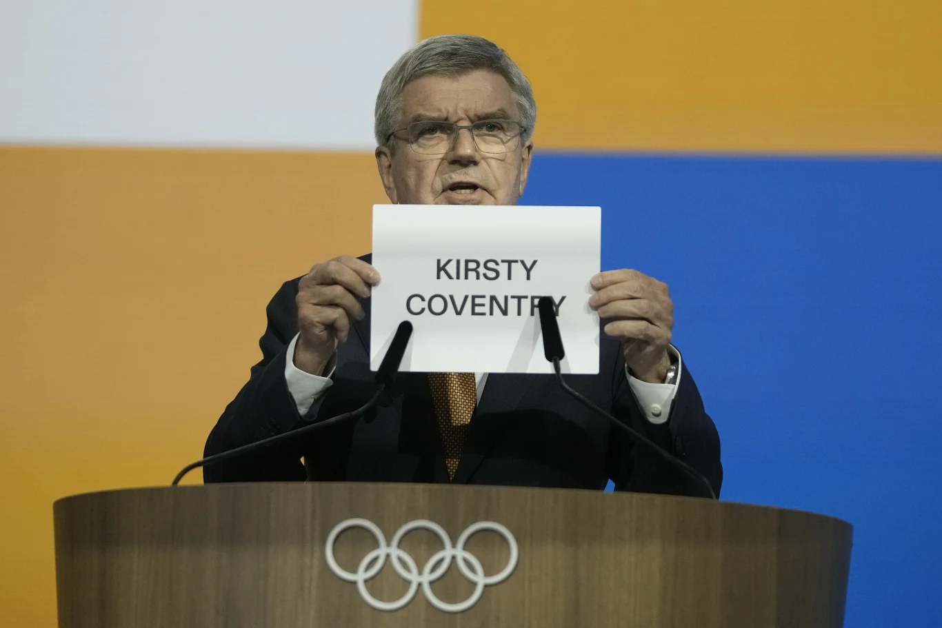 Mężczyzna w garniturze stoi za mównicą z logo olimpijskim i trzyma kartkę z napisem 'KIRSTY COVENTRY', w tle kolorowe prostokąty.