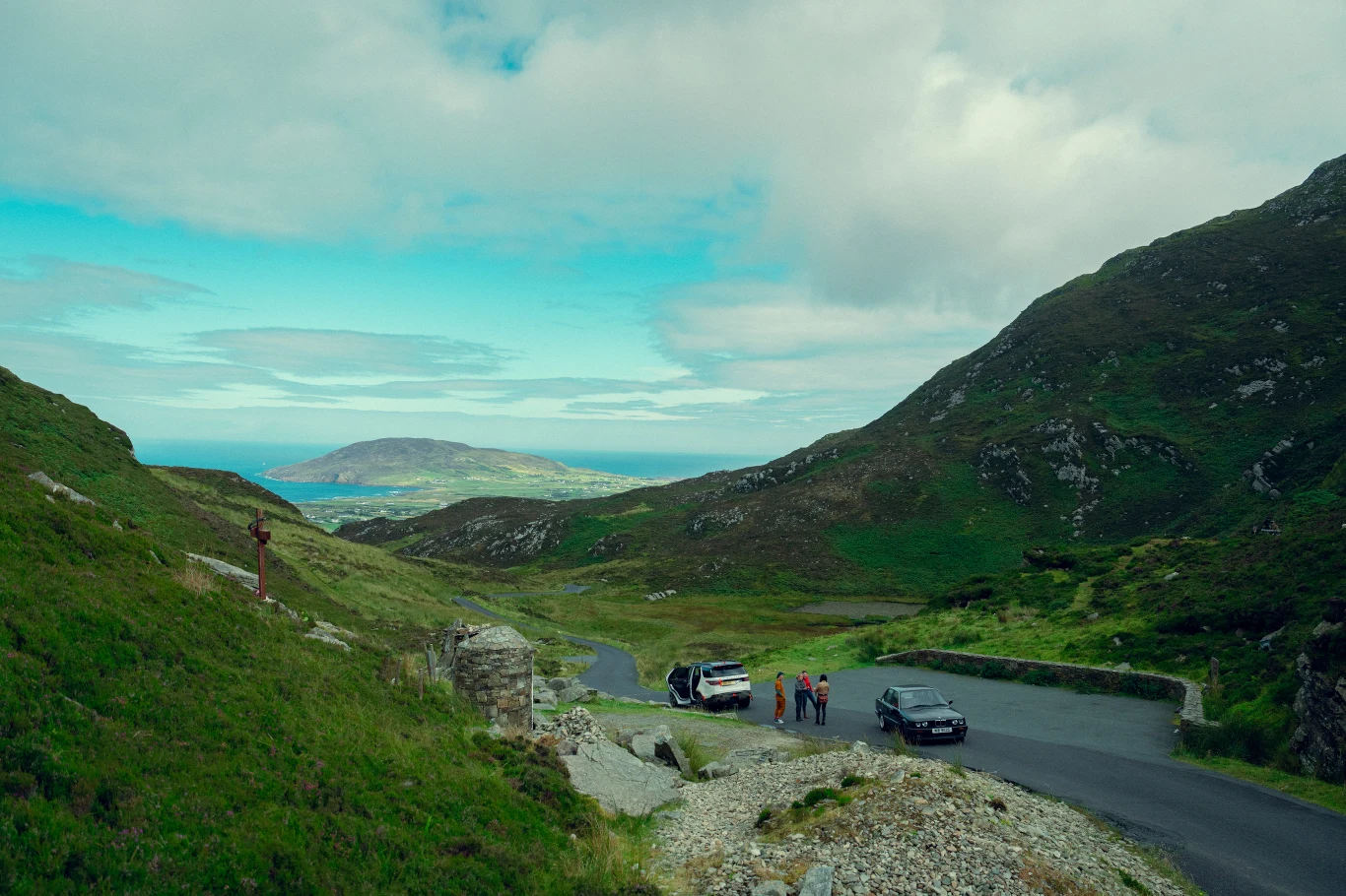 Wąska droga wśród zielonych, skalistych wzgórz, dwie zaparkowane samochody, grupa osób stojąca obok pojazdów, w oddali rozległy widok na ocean oraz wyspę pod niebieskim, lekko zachmurzonym niebem.