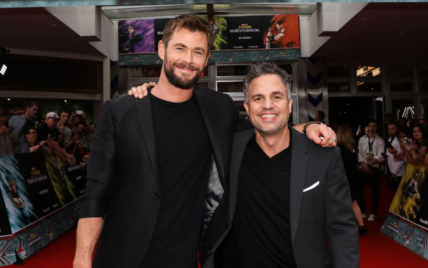 Chris Hemsworth i Mark Ruffalo Dwóch mężczyzn ubranych elegancko na czerwonym dywanie, uśmiechnięci, przytulają się ramionami, w tle tłum ludzi i plakaty filmowe.