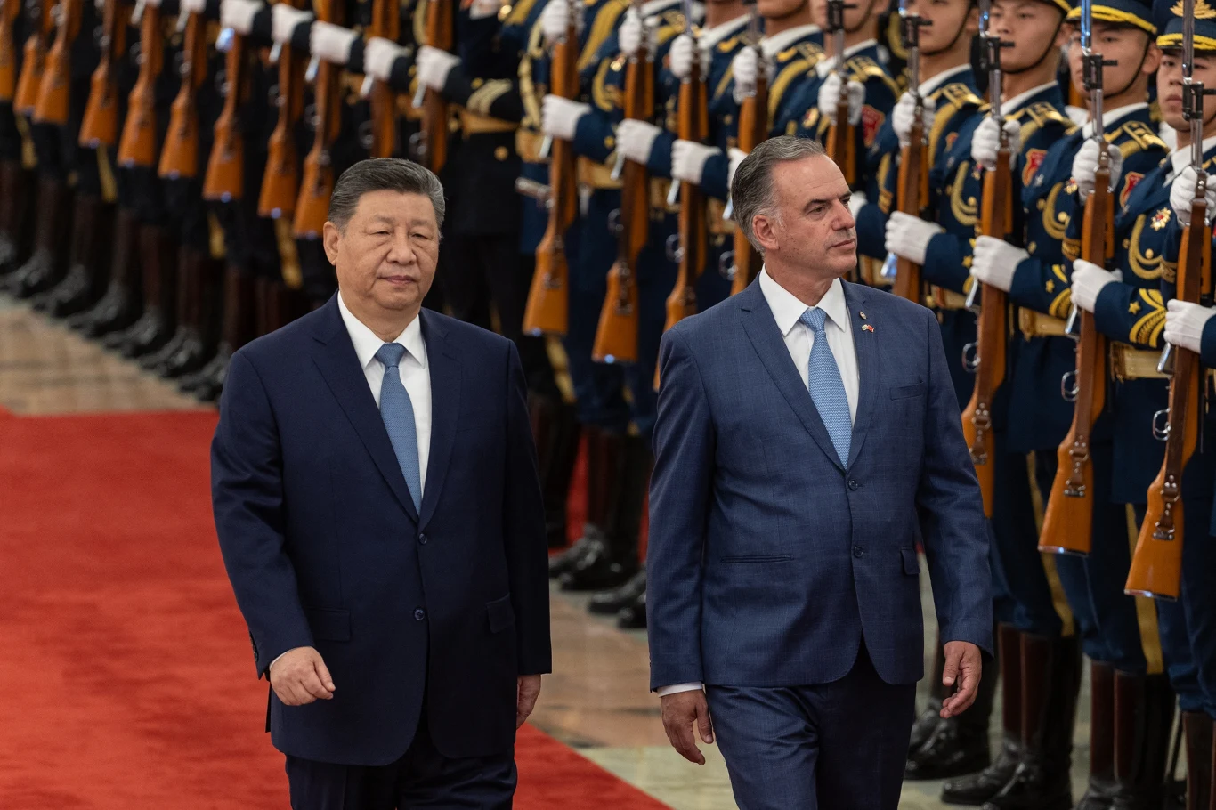 Chiński przywódca Xi Jinping oraz prezydent Urugwaju Yamandú Orsi Dwóch mężczyzn w garniturach idzie obok siebie po czerwonym dywanie, w tle rząd żołnierzy w galowych mundurach z karabinami trzymanymi pionowo.