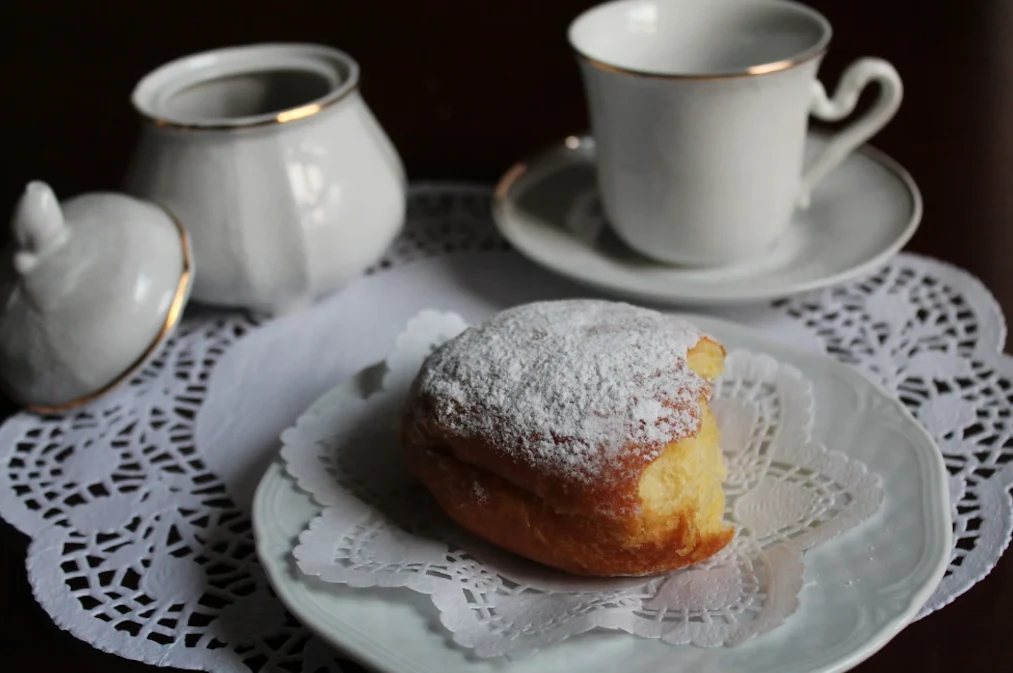 Przepis na pączki Ewy Wachowicz nie tylko na tłusty czwartek Pączek posypany cukrem pudrem na białej serwetce, obok porcelanowa filiżanka z talerzykiem oraz cukiernica z przykrywką, całość ustawiona na okrągłej serwetce.