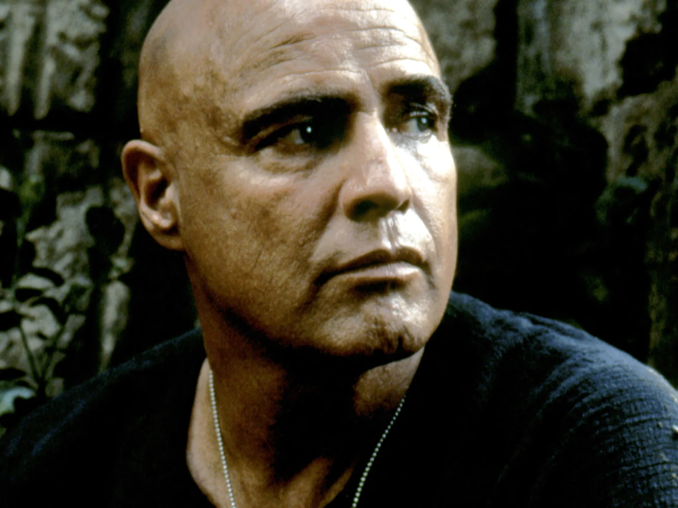 Marlon Brando w filmie "Czas apokalipsy" Łysy mężczyzna o poważnym wyrazie twarzy ubrany w ciemną koszulkę, z łańcuszkiem na szyi, siedzi na tle niewyraźnej zieleni i kamiennej ściany.