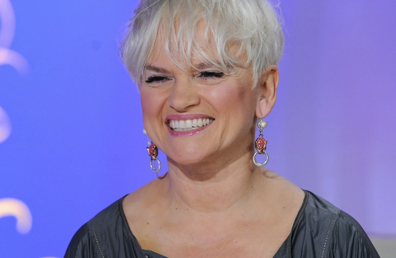Katarzyna Figura Uśmiechnięta kobieta z krótkimi, białymi włosami w eleganckiej sukience, nosząca długie ozdobne kolczyki na tle fioletowego światła.
