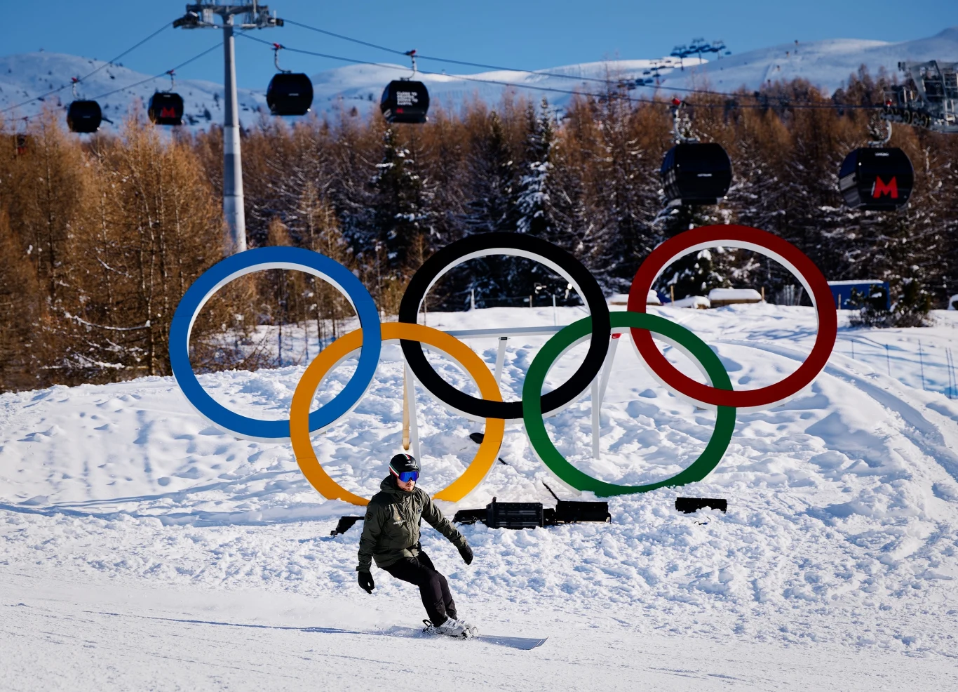 Snowboardzista sunie pośnieżnej trasie na tle dużych, kolorowych kół olimpijskich, w tle widać drzewa oraz ośnieżone góry i kursujące gondole.